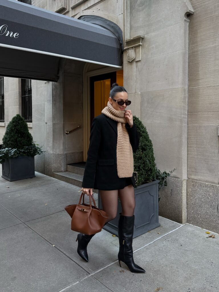 Woman in a black tailored coat with a chunky camel knit scarf, black mini skirt, sheer tights, black knee boots and a brown tote
