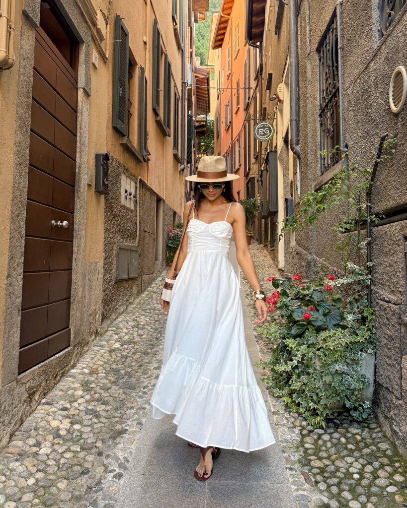 Woman wearing a flowy white tiered maxi dress with a straw fedora and brown sandals in a cobblestone alley in Bellagio, Italy