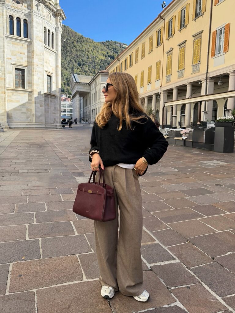 Woman wearing a black crewneck sweater, taupe wide-leg trousers, and white sneakers holding a burgundy handbag on a piazza in Como, Italy