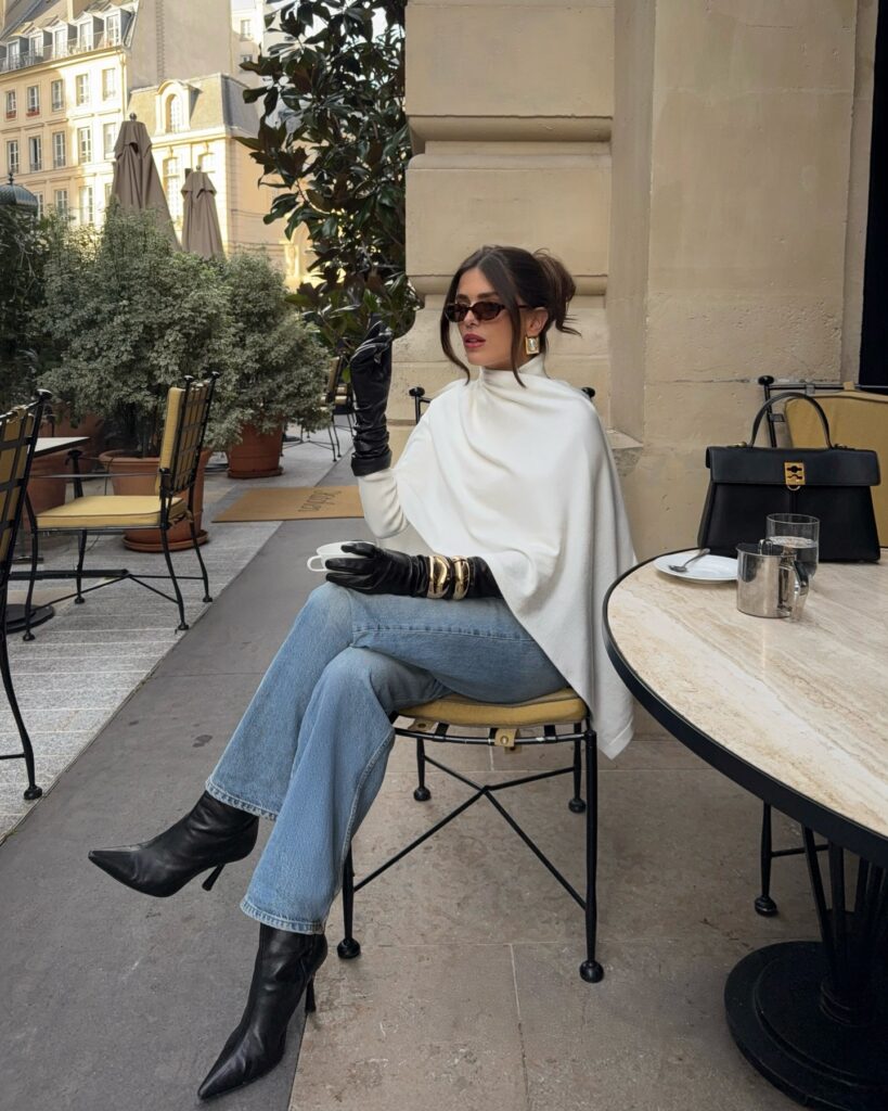 Woman seated at a café terrace wearing a white cape over a knit top, long black leather gloves with gold bangles, blue jeans, and black heeled boots