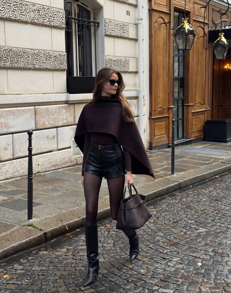 Woman wearing a chocolate brown cape over black leather shorts, sheer tights, and black knee-high boots carrying a structured brown tote on a cobblestone street