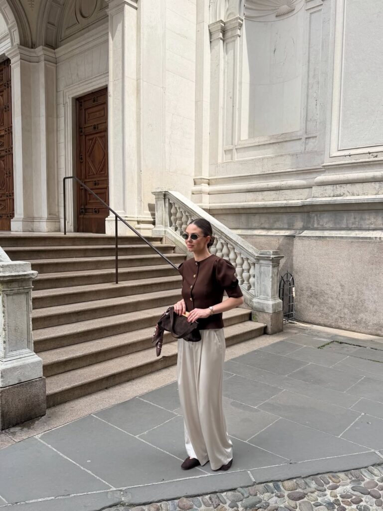 Woman wearing a chocolate brown puff-sleeve button top and cream satin wide-leg trousers with brown flats standing near marble steps in Italy
