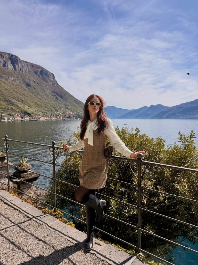 Woman wearing a brown plaid mini dress layered over a white bow blouse with black tights and black knee-high boots by a railing overlooking Lake Como