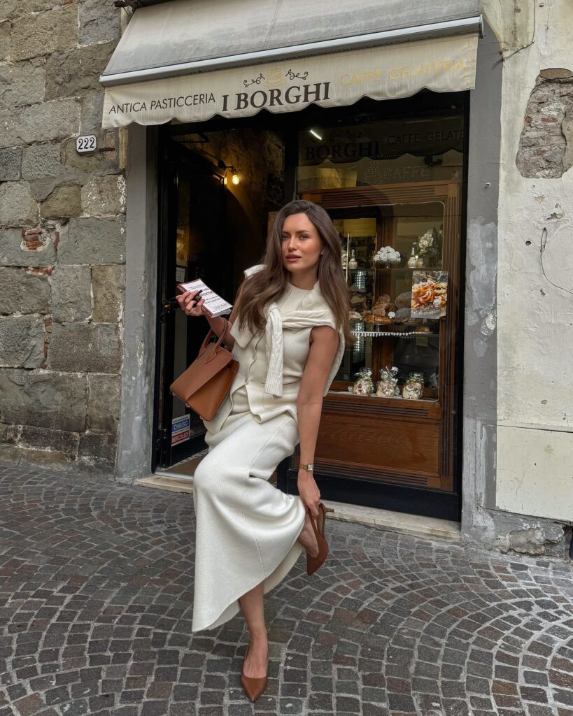 Woman wearing a cream knit vest and matching knit maxi skirt with a sweater draped over her shoulders and tan slingback heels outside an Italian pasticceria