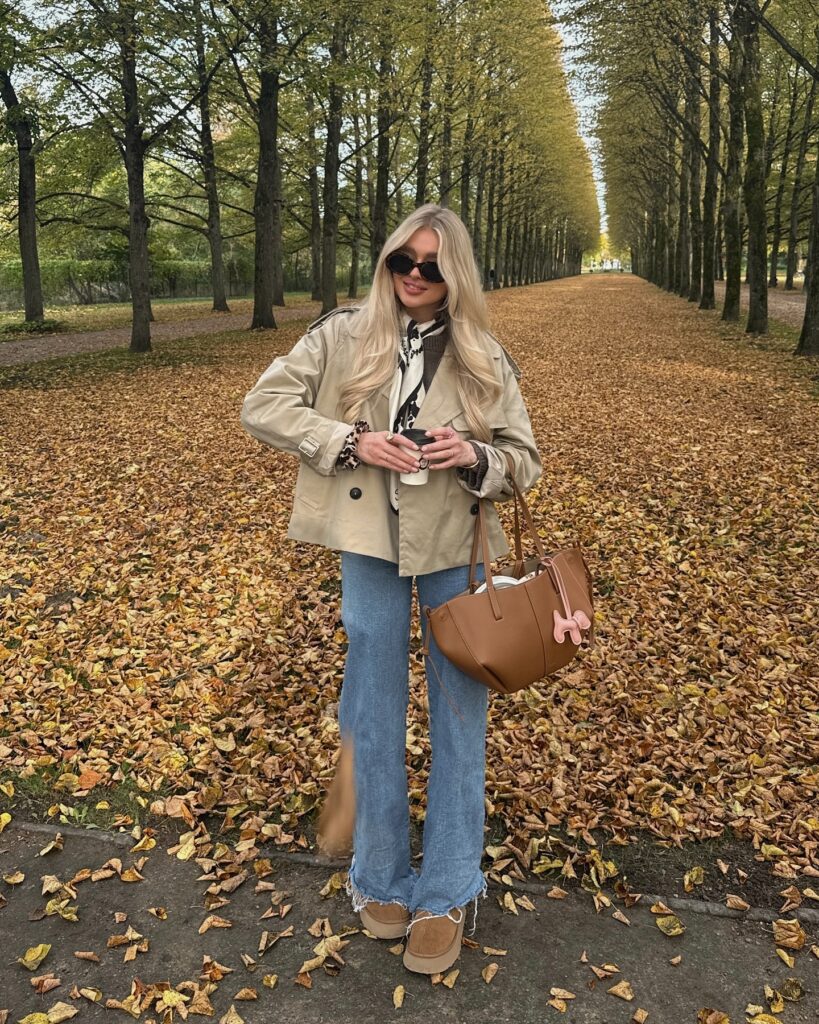 Woman in a cropped beige trench coat, printed scarf, flared jeans, and tan platform boots standing on a tree-lined path covered in autumn leaves