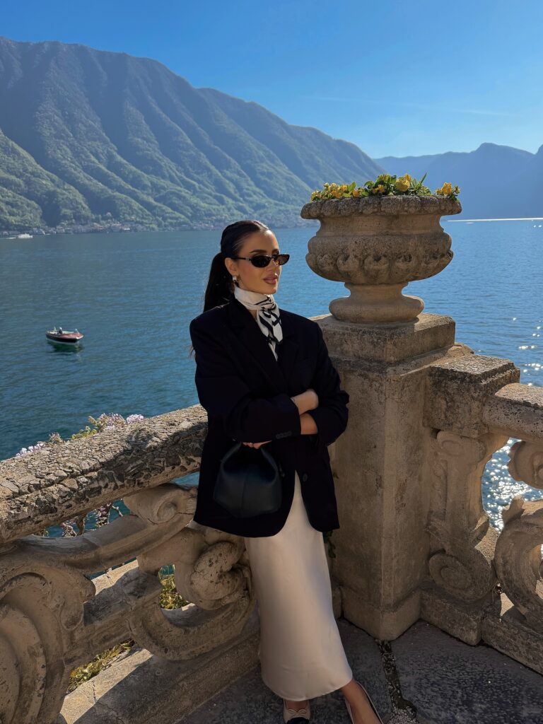 Woman wearing an oversized navy blazer, white and black silk neck scarf, and cream satin maxi skirt leaning against a stone balustrade overlooking Lake Como