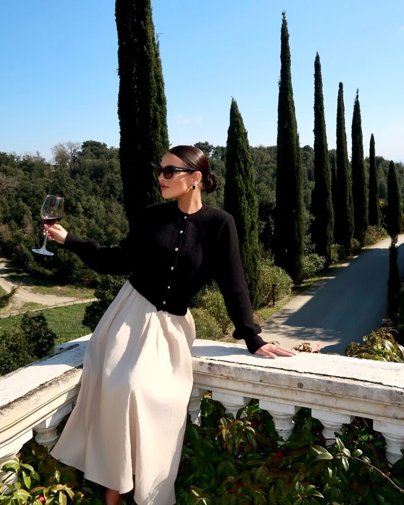 Woman holding a glass of red wine wearing a black jewel-button cardigan and cream midi skirt on a white balustrade terrace lined with cypress trees in Tuscany