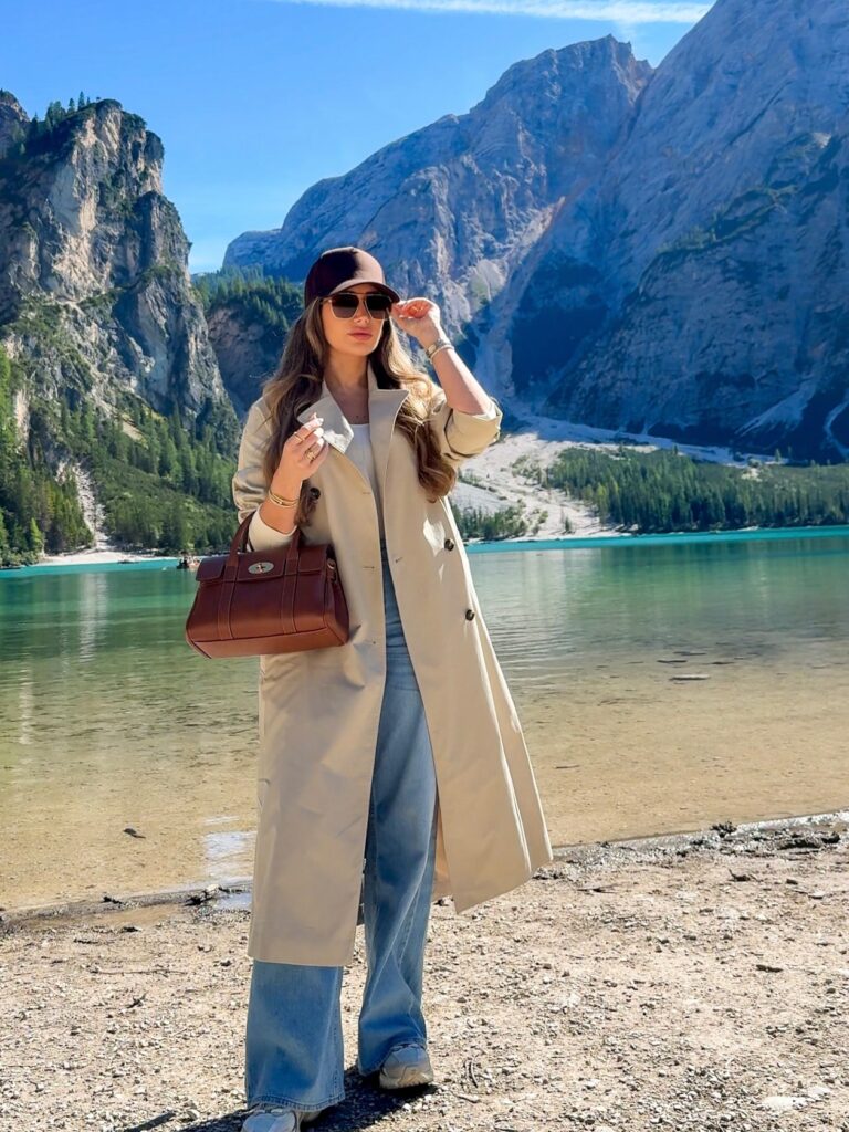 Woman wearing a stone trench coat, brown baseball cap, wide-leg jeans, and sneakers with a brown leather bag at Lago di Braies in the Italian Dolomites