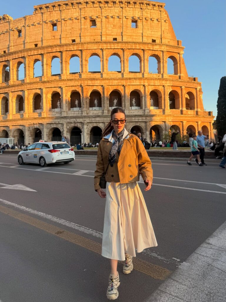 Woman wearing a cropped tan trench jacket, navy bandana scarf, cream pleated midi skirt, and sneakers in front of the Colosseum at sunset in Rome