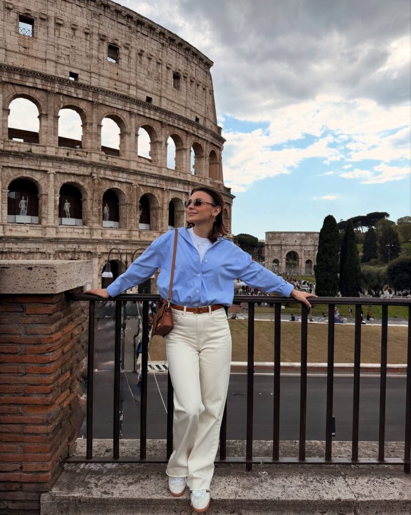 Woman in an oversized light blue button-down shirt, cream wide-leg jeans with a tan belt, and white sneakers leaning on a railing in front of the Colosseum in Rome