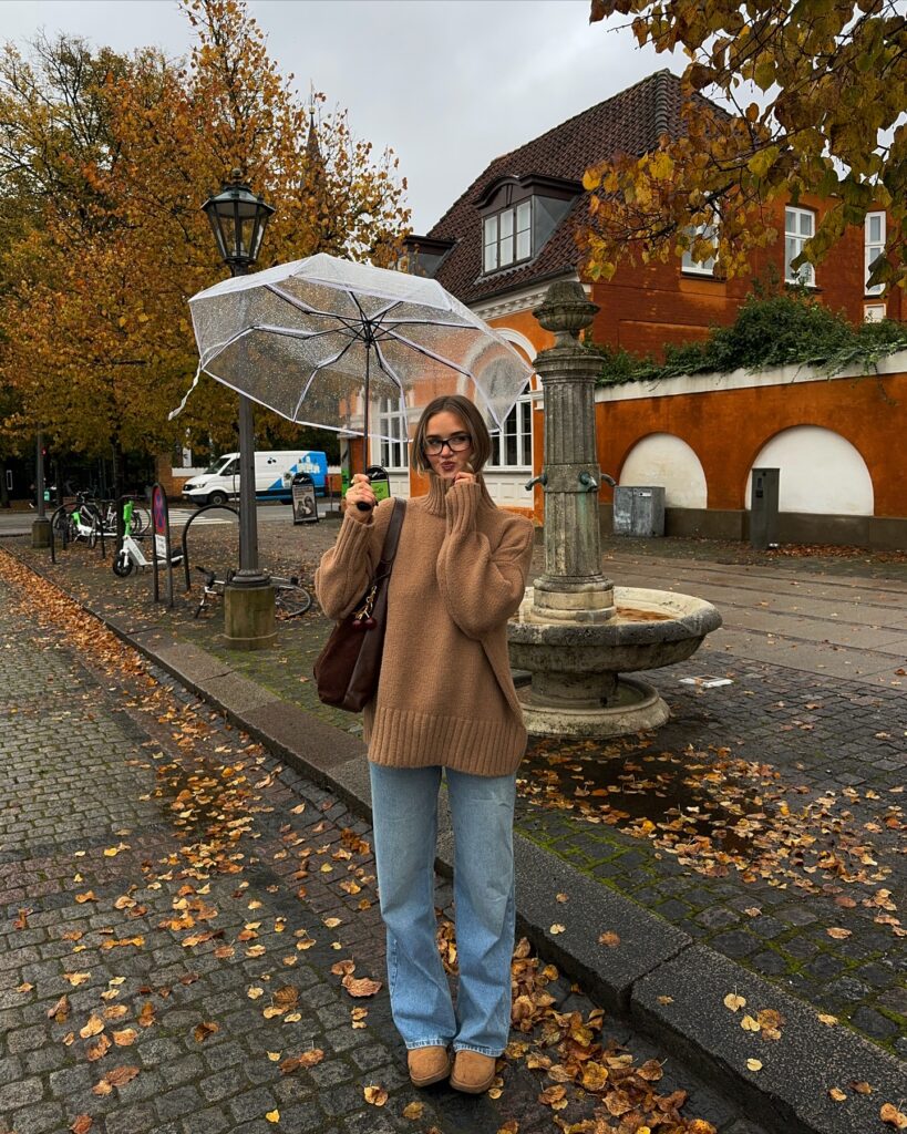 Woman holding a clear umbrella wearing an oversized camel turtleneck sweater, light blue jeans, and tan suede boots on a rainy fall day