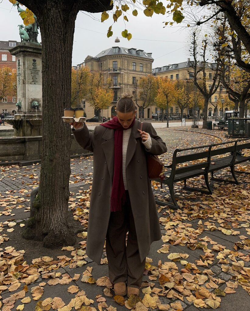 Woman carrying two coffees wearing a long taupe wool coat, burgundy scarf, cream knit sweater, and brown trousers in a leaf-covered square