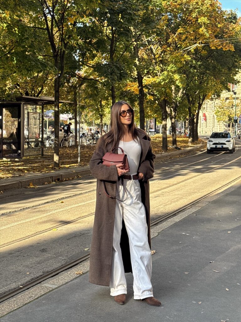 Woman in a long brown wool maxi coat over a white top and white wide-leg jeans with brown suede boots, standing on a tram-lined street in Milan in autumn