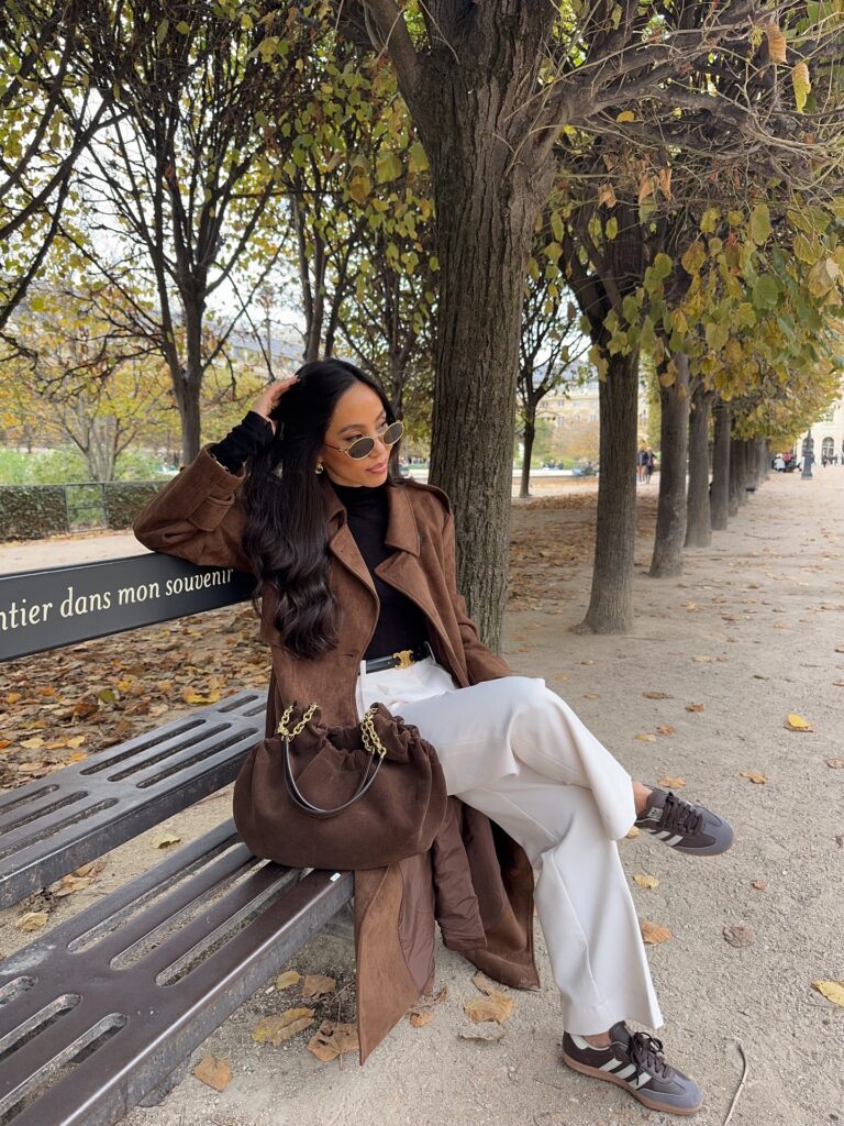 Woman sitting on a Paris park bench in autumn wearing a brown suede trench coat over a black top, white wide-leg trousers, and brown retro sneakers with a brown suede bag