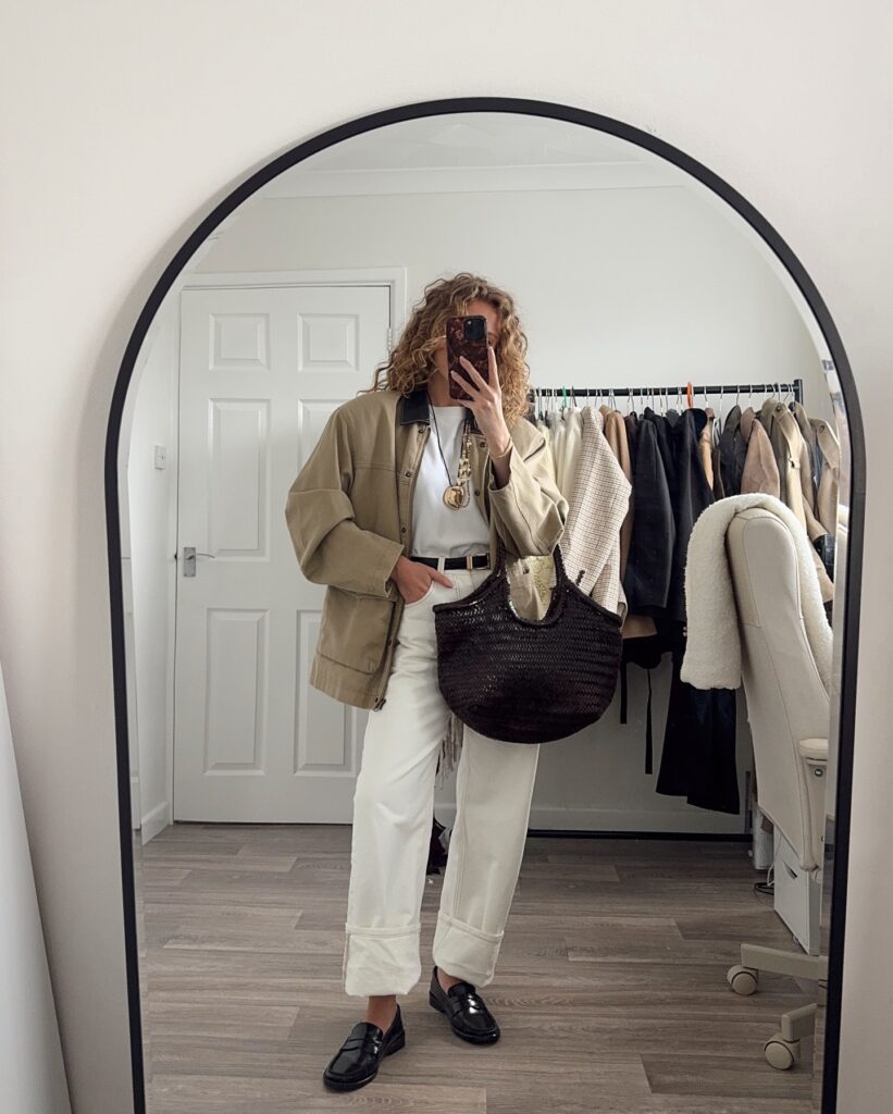 Mirror selfie of a woman wearing a khaki utility chore jacket over a white tee, cream cuffed jeans, black penny loafers, and a woven brown tote bag