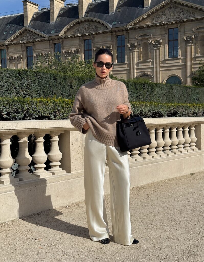 Woman standing in front of a classic Paris building wearing a taupe chunky turtleneck sweater, ivory satin wide-leg trousers, black cap-toe flats, and a black handbag
