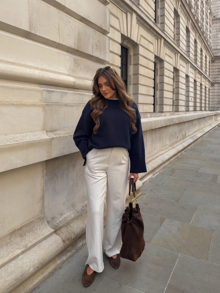Woman leaning against a stone building wearing an oversized navy sweater, cream tailored trousers, brown suede flats, and a brown suede bag