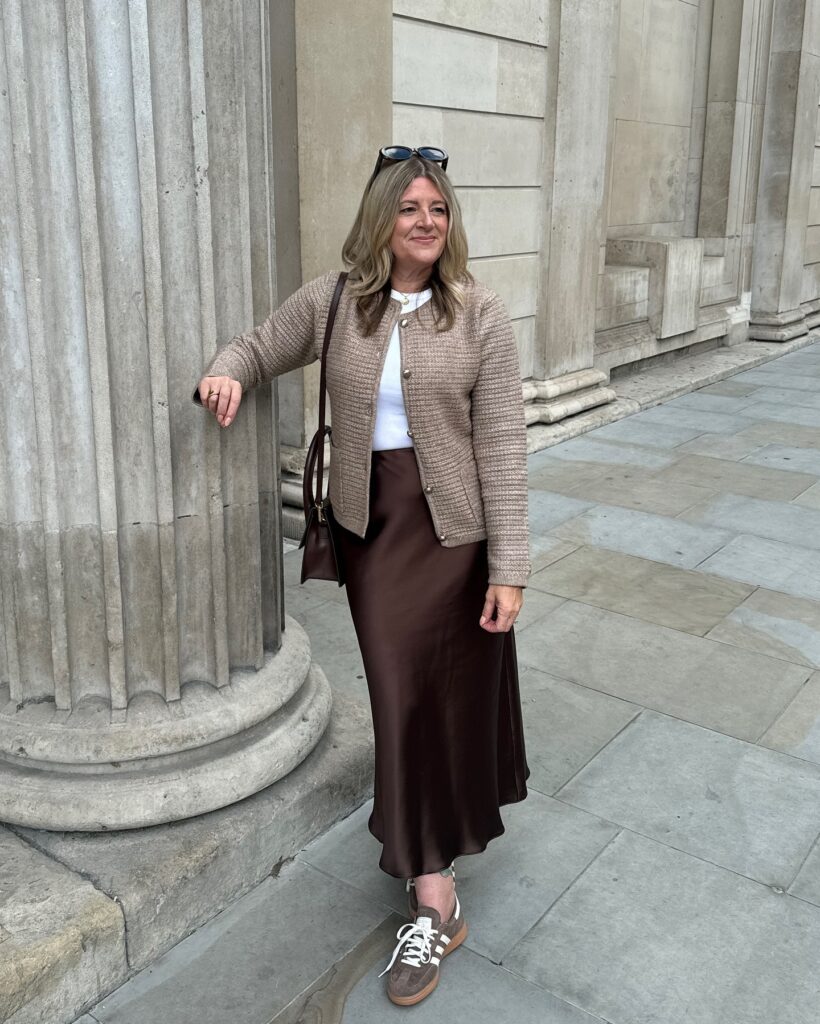 Woman leaning against a stone column wearing a textured taupe cardigan jacket over a white top, chocolate brown satin midi skirt, and brown suede sneakers