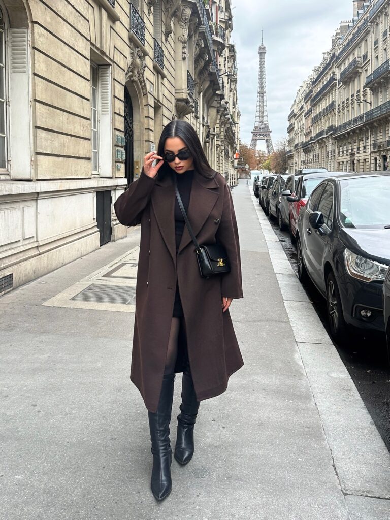Woman sitting on a green park bench in fall wearing a grey chunky turtleneck under an oversized black blazer with sheer tights and tall black leather boots, holding a brown suede bag