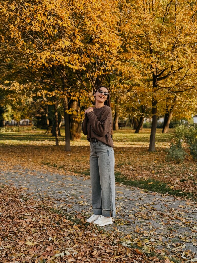 Woman standing under golden autumn trees wearing a brown oversized crewneck sweater, light wash wide-leg jeans, and white sneakers