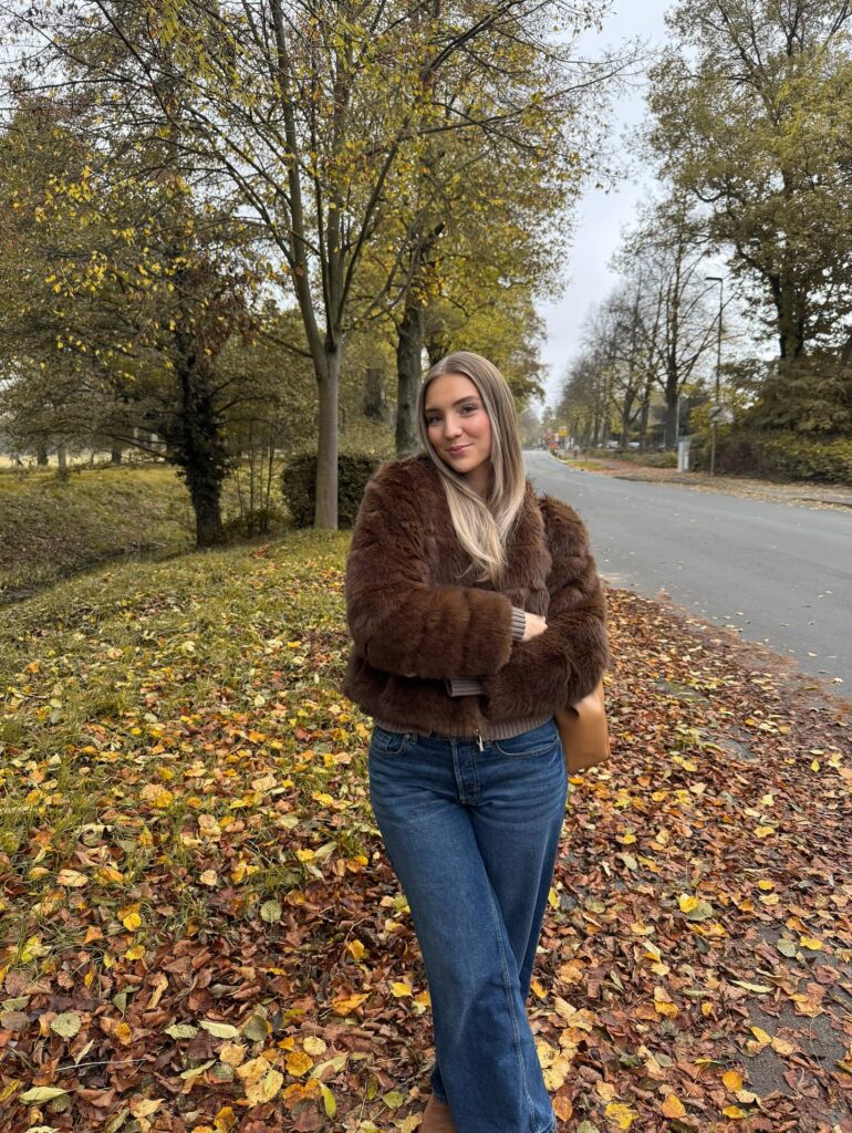 Woman standing on a leafy autumn roadside wearing a chocolate brown faux fur jacket, medium wash straight jeans, and a tan shoulder bag