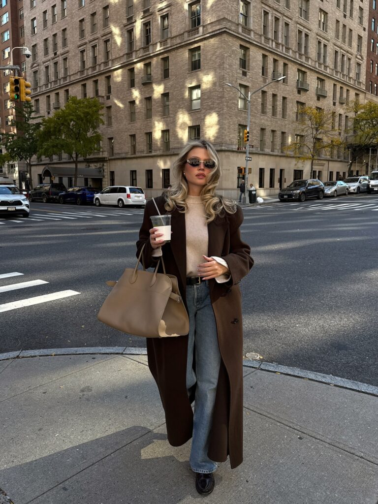 Woman crossing a New York street wearing a long chocolate brown wool coat over a beige sweater, light wash jeans, dark loafers, and a taupe tote bag
