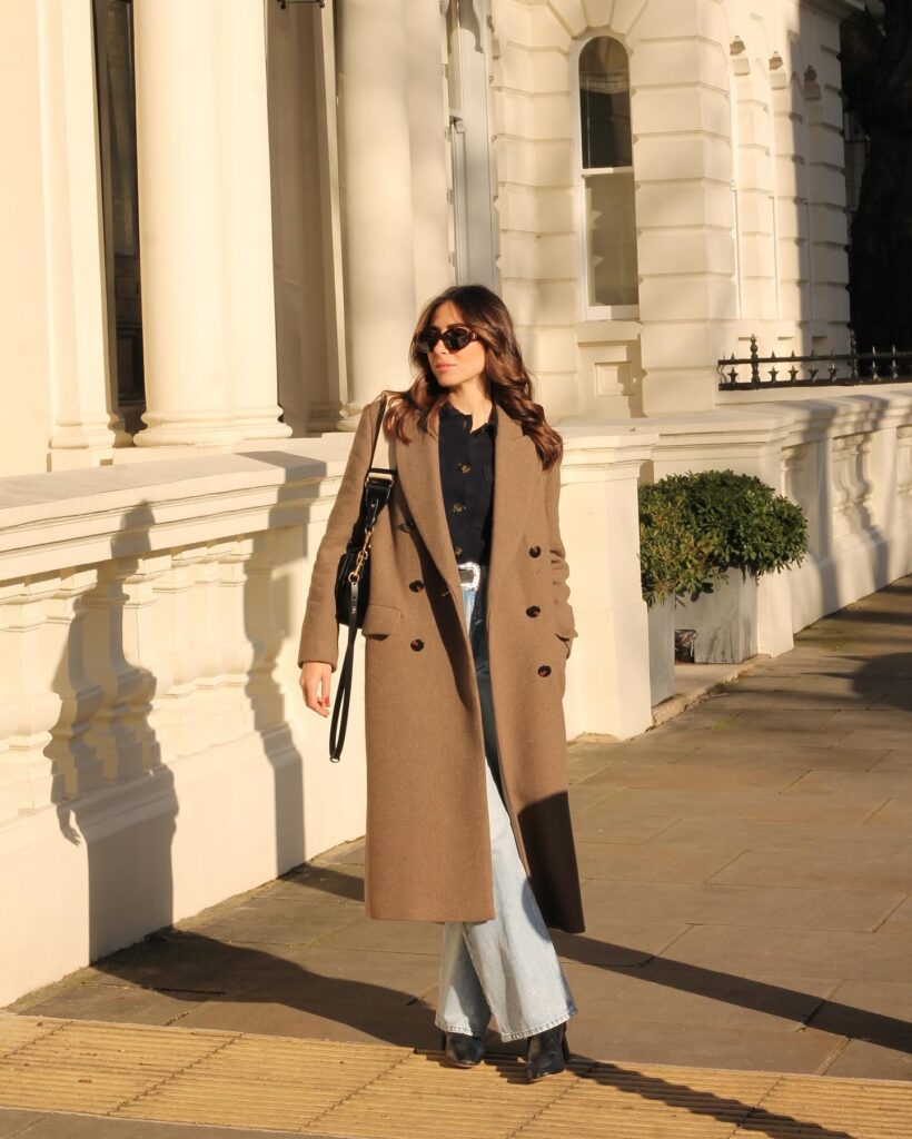Woman in golden sunlight wearing a long camel wool coat over a navy shirt, light wash wide-leg jeans, and black boots with a black shoulder bag