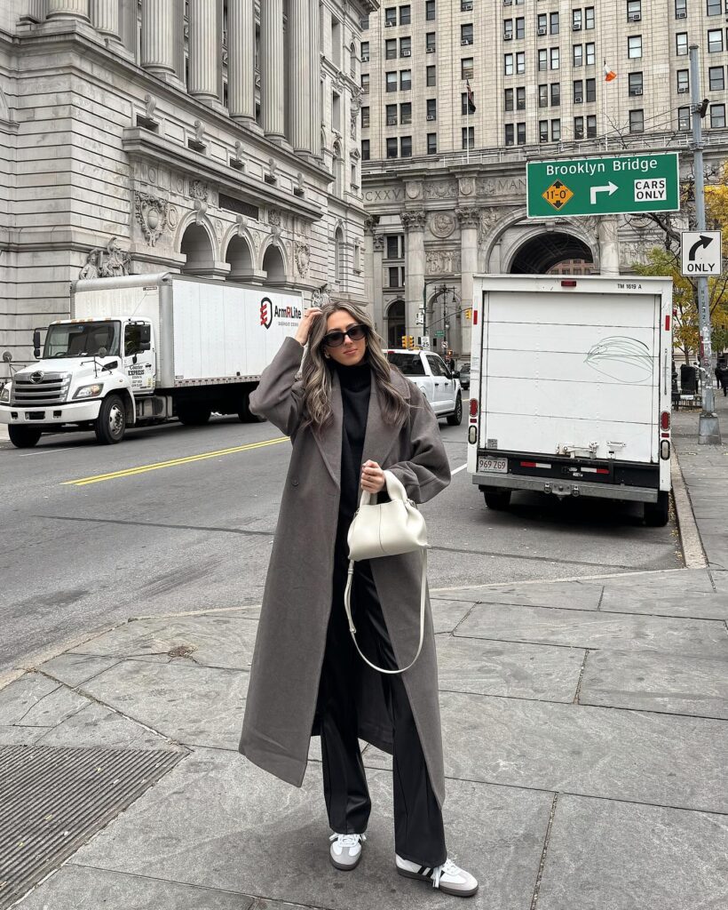 Woman on a New York City street wearing a long grey wool coat over a black turtleneck and black trousers with white sneakers and a small cream bag