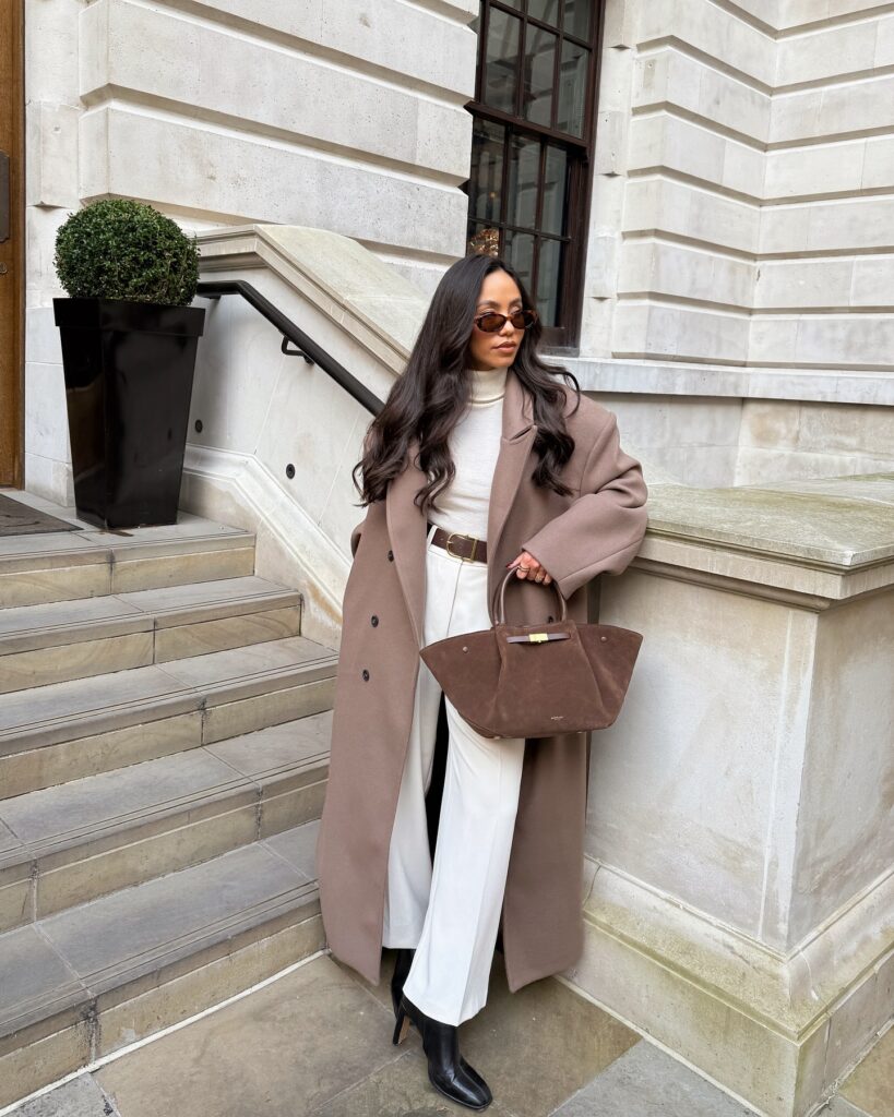 Woman on stone steps wearing an oversized taupe wool coat over a cream turtleneck and cream trousers with a brown belt, black heeled boots, and a brown suede bag