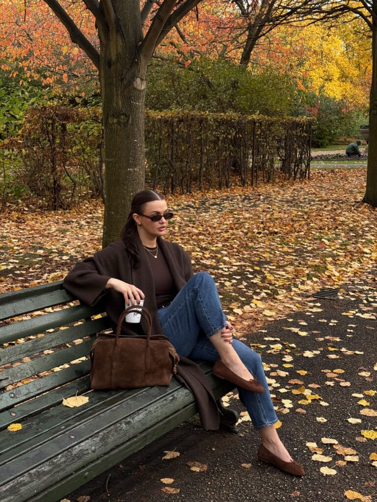 Woman on stone steps wearing an oversized taupe wool coat over a cream turtleneck and cream trousers with a brown belt, black heeled boots, and a brown suede bag
