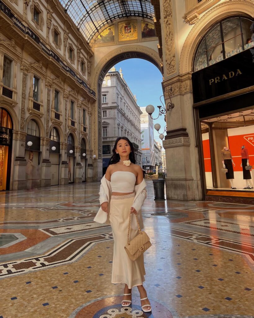 Woman in a white bandeau top, cream satin midi skirt, white blazer, and rosette choker inside the Galleria Vittorio Emanuele II in Milan