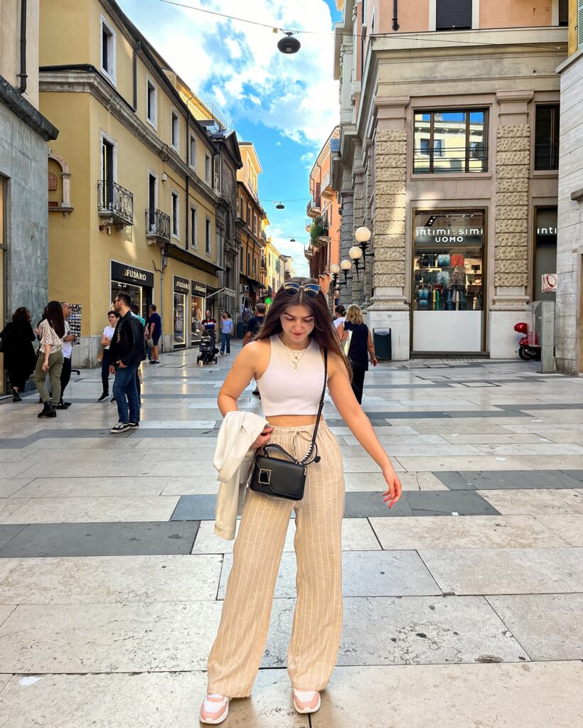 Woman wearing a white racerback crop top and striped beige linen drawstring pants with sneakers in a shopping street in Verona