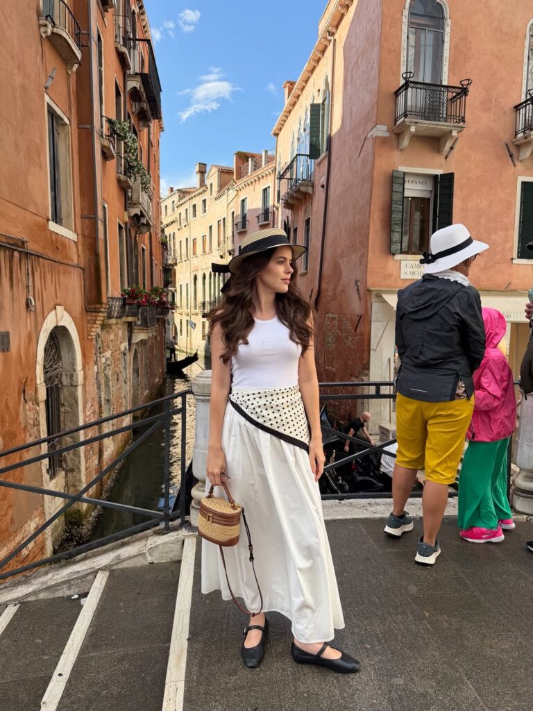 Woman wearing a white tank, white maxi skirt with a polka dot scarf tied at the hip, boater hat, and black Mary Jane flats on a Venice bridge