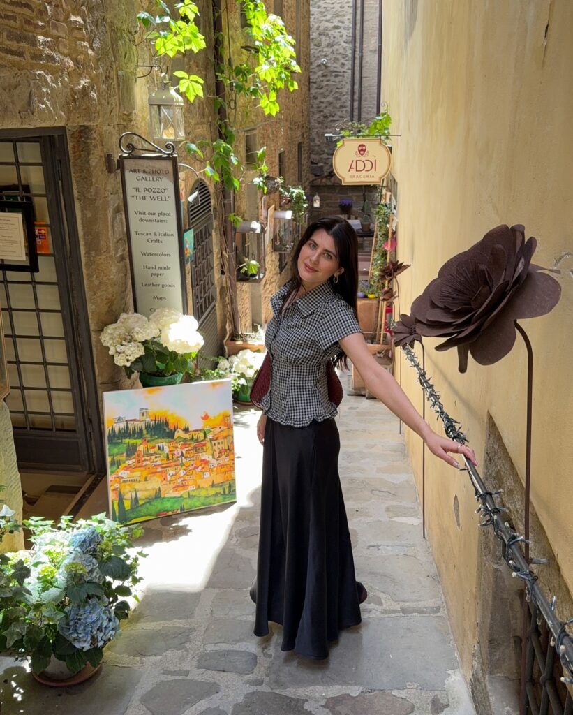Woman in a black and white gingham short-sleeve blouse and long black satin skirt in a narrow flower-lined alley in Cortona, Tuscany