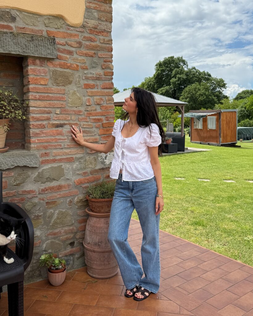 Woman wearing a white eyelet puff-sleeve tie-front blouse with relaxed light-wash jeans and dark sandals at a stone Tuscan farmhouse