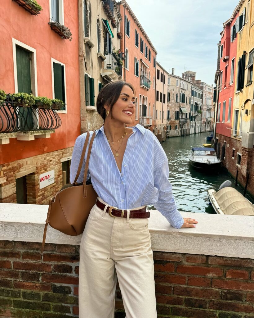 Woman in an oversized blue striped button-down tucked into cream jeans with a brown croc belt and tan tote on a Venice canal bridge