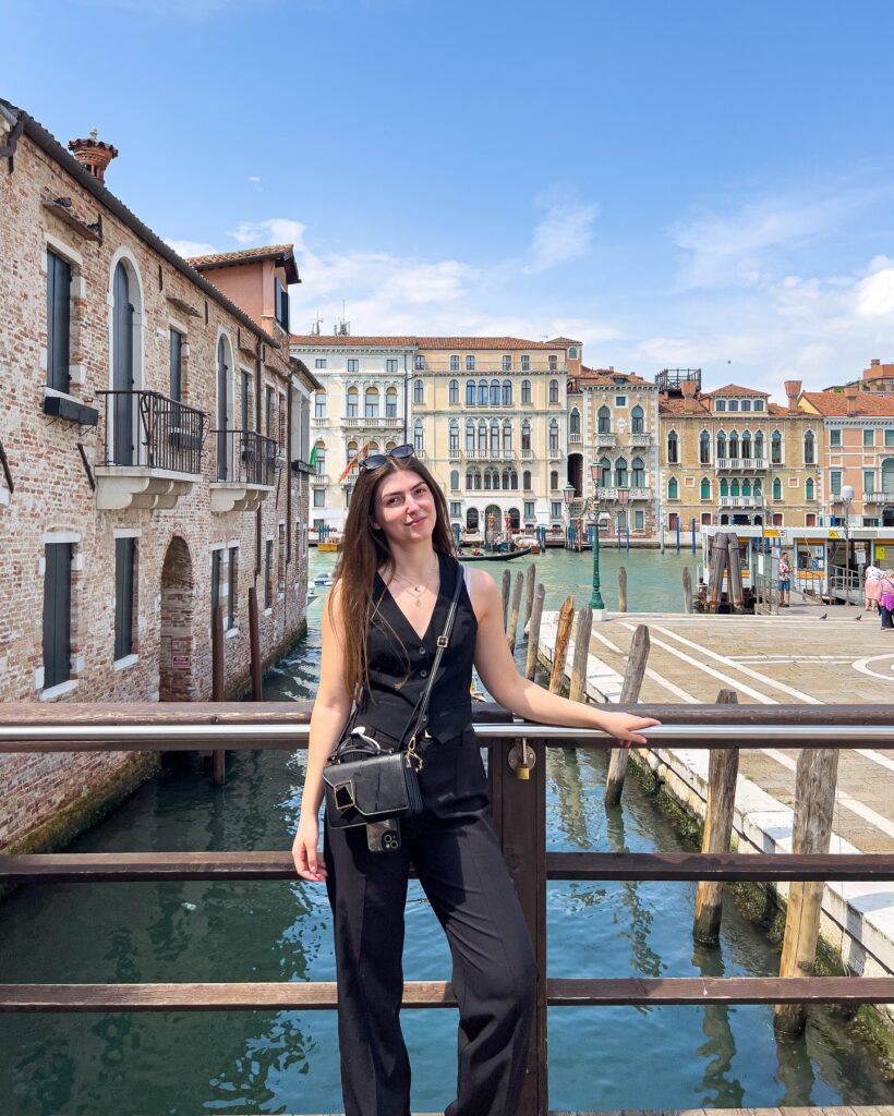 Woman wearing a black waistcoat vest and black trousers with a small crossbody bag on a bridge over the Grand Canal in Venice