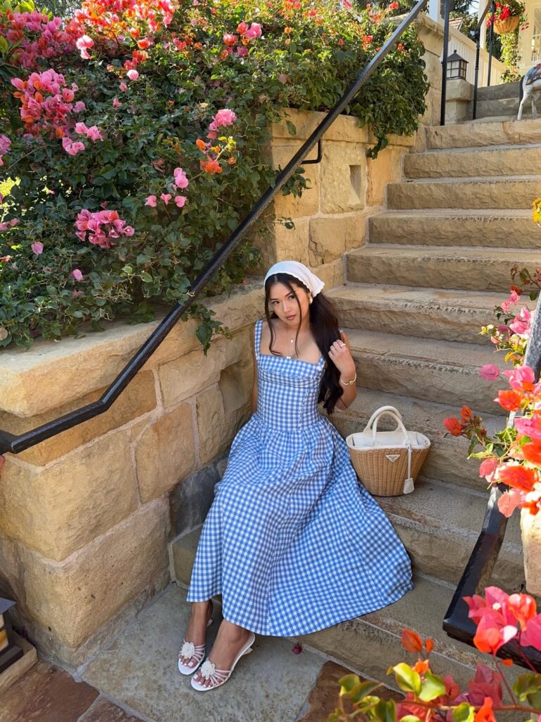 Woman in a blue and white gingham corset-bodice midi dress with a white headscarf and wicker bag sitting on stone steps beside bougainvillea