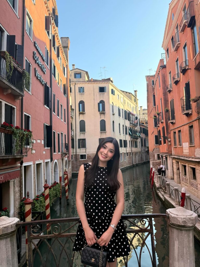 Woman wearing a black sleeveless polka dot mini dress with a quilted black chain bag standing on a small bridge over a Venice canal