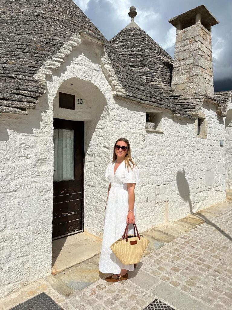 Woman wearing a white flutter-sleeve maxi dress with a straw tote and suede Mary Janes in front of the trulli houses of Alberobello, Puglia