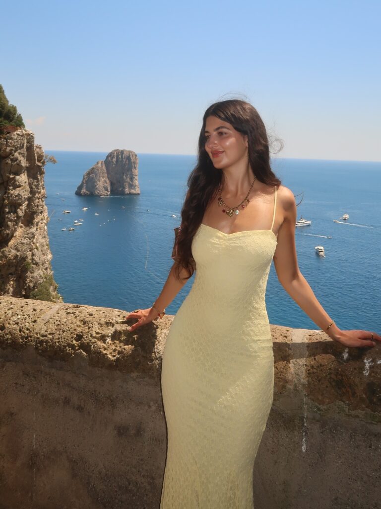 Woman wearing a pale yellow lace slip maxi dress with a charm necklace overlooking the Faraglioni rocks in Capri