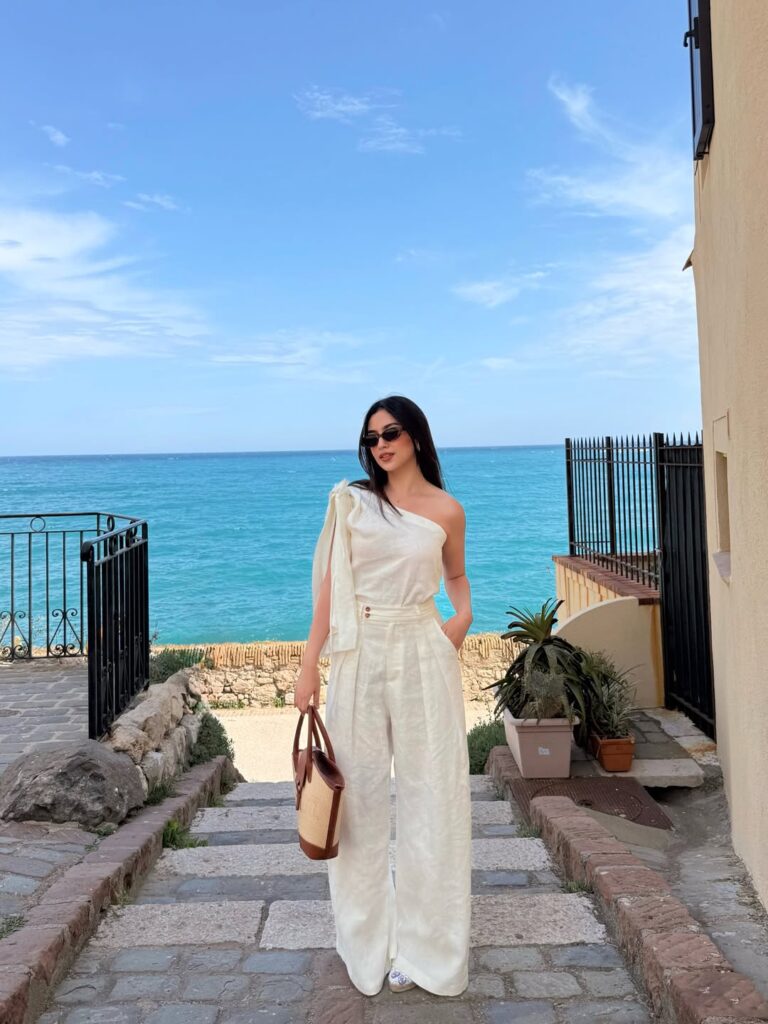 Woman wearing a white one-shoulder linen top with matching pleated wide-leg trousers and a straw tote by the turquoise sea