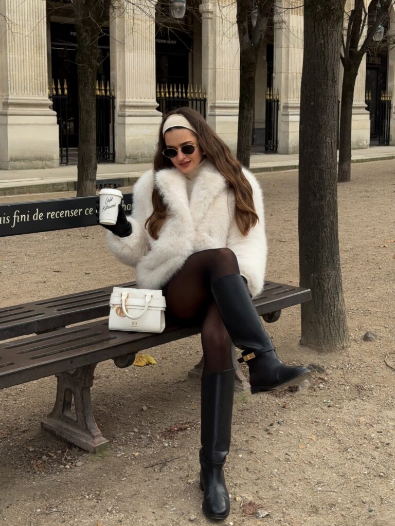 Woman sitting on a park bench at Palais Royal in Paris wearing a white faux fur jacket, black tights, black knee-high riding boots, a cream headband, and holding a Café Kitsuné coffee cup.