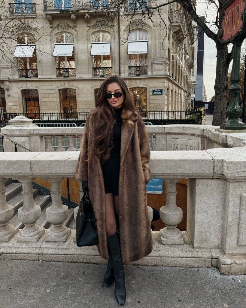 Woman standing by a Paris Métro entrance wearing a long brown faux fur coat over a black turtleneck and black shorts with black knee-high boots and a black handbag.