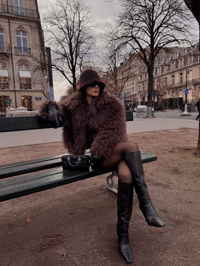 Woman sitting on a green Parisian park bench in winter wearing a shaggy brown teddy coat, matching fuzzy bucket hat, black tights, leather gloves, and black knee-high boots with a quilted black bag.