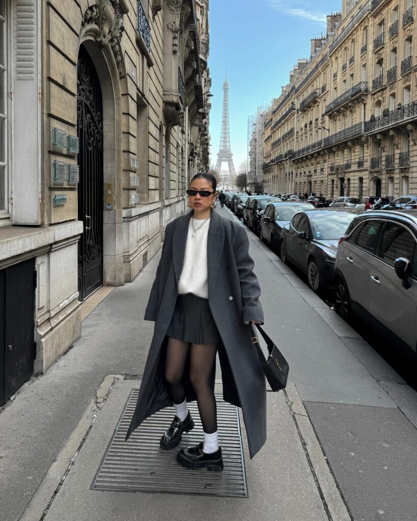 Woman standing on a Paris sidewalk with the Eiffel Tower at the end of the street, wearing a long oversized grey coat, white fuzzy sweater, grey pleated mini skirt, black tights, white socks, and chunky black loafers.