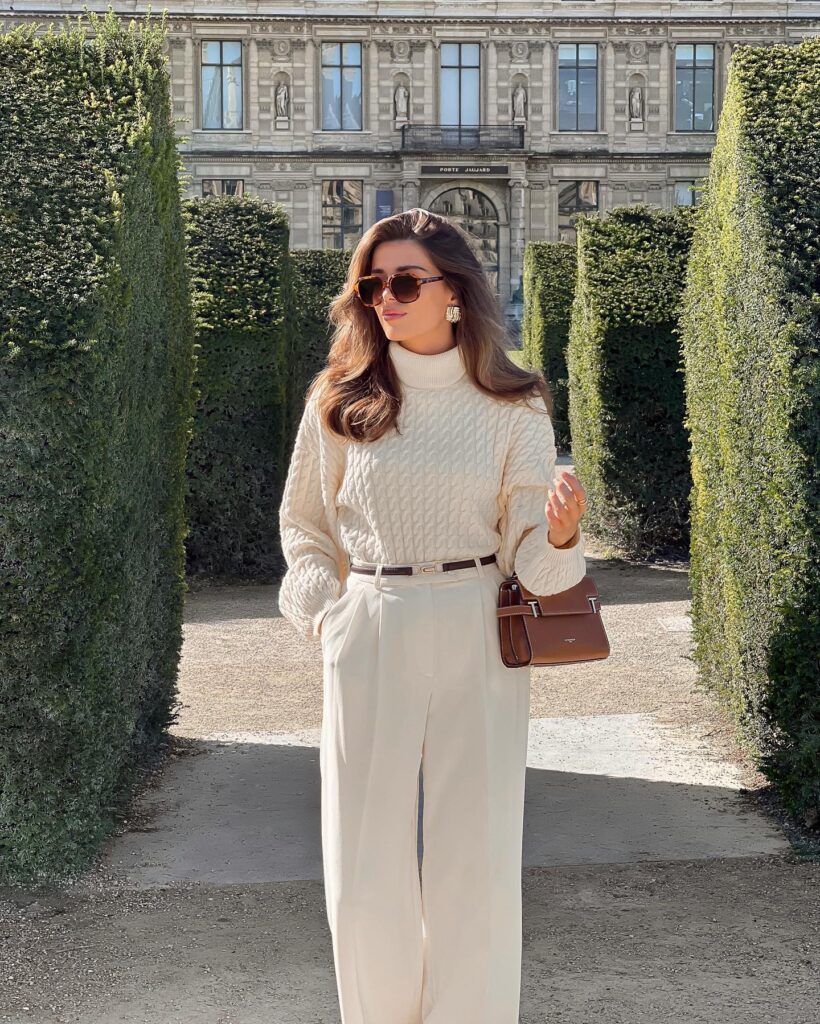 Woman standing between tall green hedges in the Tuileries Garden in Paris wearing a cream cable-knit turtleneck sweater tucked into cream wide-leg trousers with a slim brown belt, tortoiseshell sunglasses, and a small brown handbag.