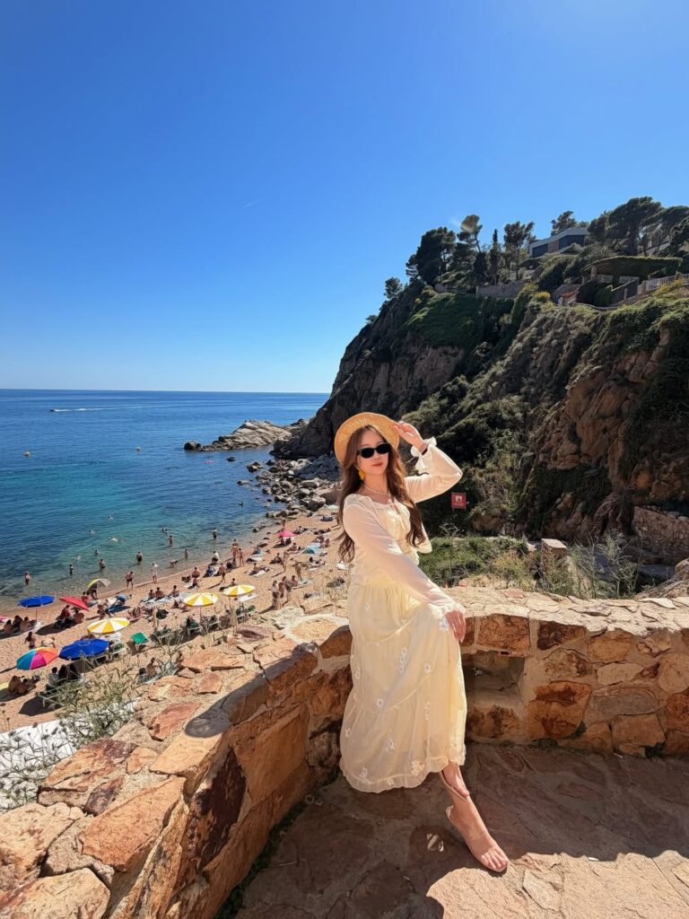 Woman wearing a pale yellow long-sleeve maxi dress and straw boater hat overlooking a busy beach cove on the Costa Brava, Spain