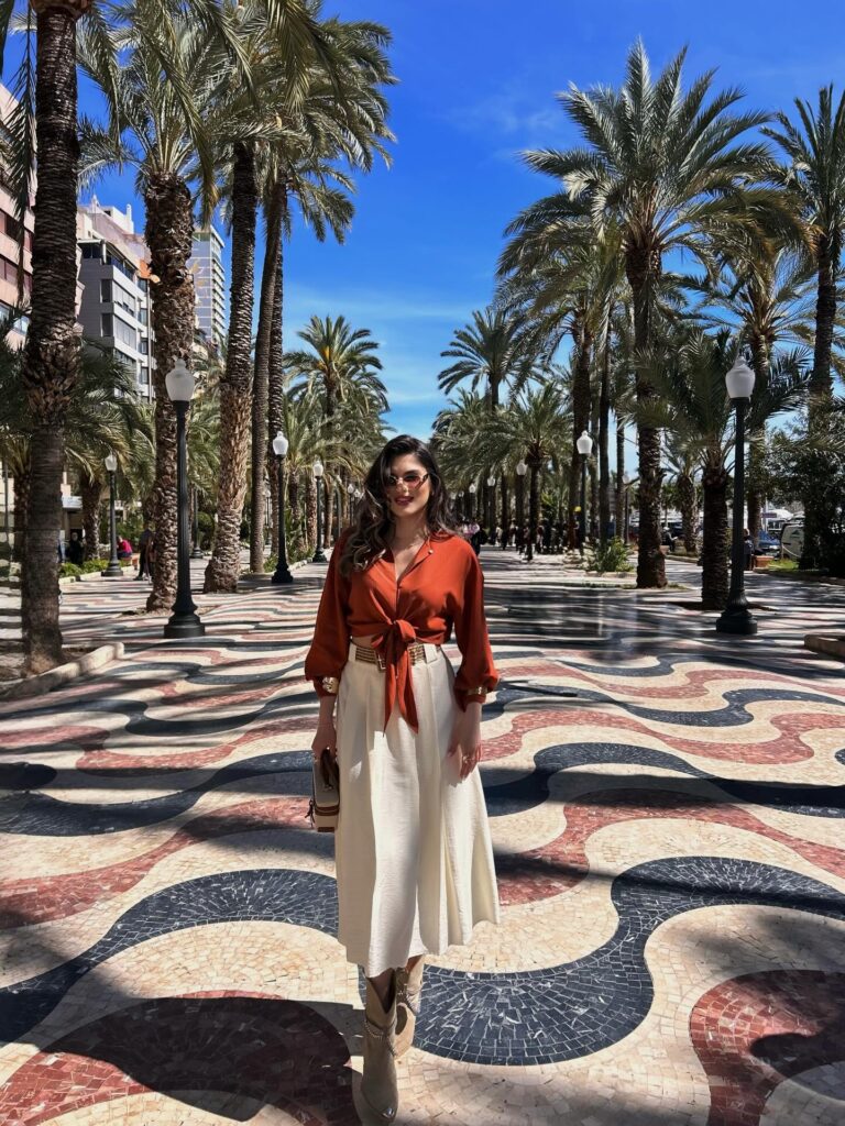 Woman in a rust orange tie-front blouse and cream pleated midi skirt walking the wavy mosaic Explanada de España promenade in Alicante, Spain