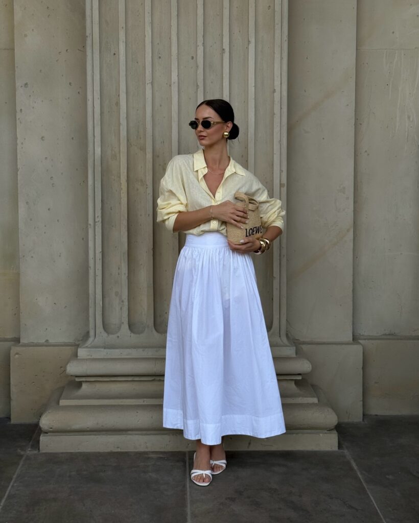 Woman in a pale butter yellow linen shirt and white full maxi skirt with white heeled sandals and a small raffia bag, standing before carved stone columns